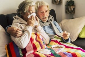 senior-couple-sit-on-sofa-woman-blows-nose-while-man-comforts-her